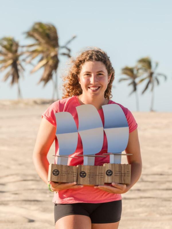 Una mujer sostiene dos cajas de Wingfoil en la playa, con palmeras en el fondo. GWA World Tour Logo en la esquina inferior izquierda.