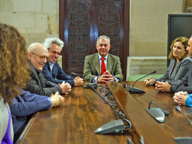 Reunión de trabajo en una sala histórica con participantes sentados alrededor de una mesa de madera con micrófonos.