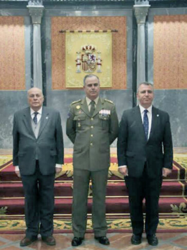 Tres individuos en trajes formales y militar posando en un salón con decoración clásica y escudo de armas en el fondo.