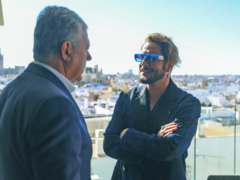 Dos hombres en una terraza con vistas a la ciudad, conversando con el sol brillando en el fondo.