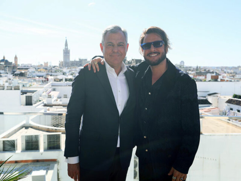 Dos hombres sonrientes posan juntos en un balcón con vistas a la ciudad de Sevilla, España.