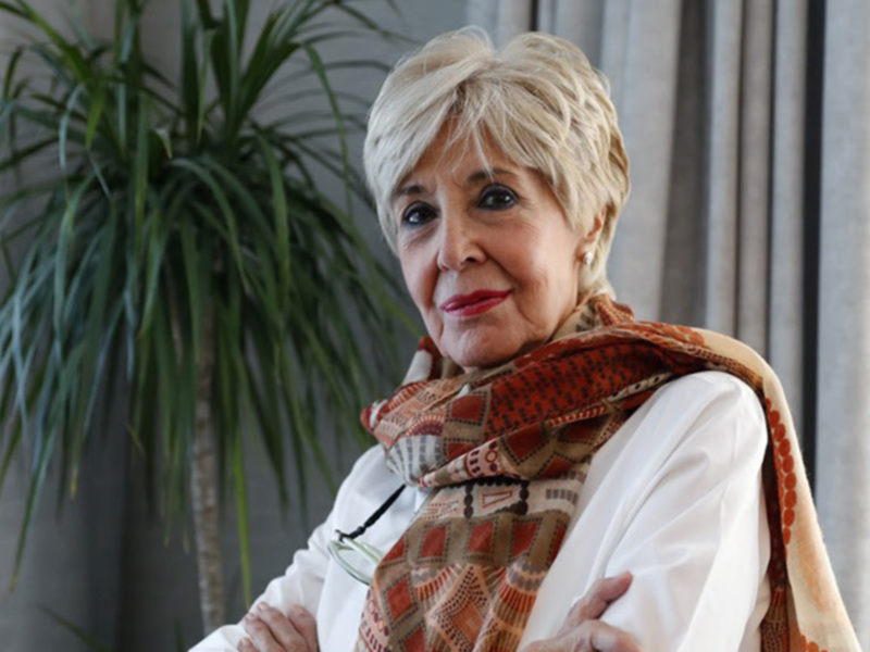 Una mujer con una bufanda de colores en un entorno interior con una planta en el fondo.