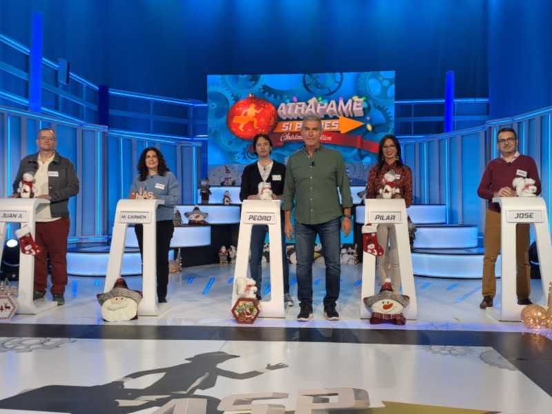 Un grupo de personas en un escenario de televisión, posiblemente en un programa de juegos o concursos, con stands individuales y decoraciones navideñas.