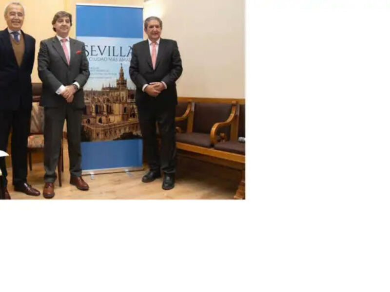 Tres hombres en traje posan frente a un cartel de 'Sevilla, Ciudad más Amable' en un ambiente formal.