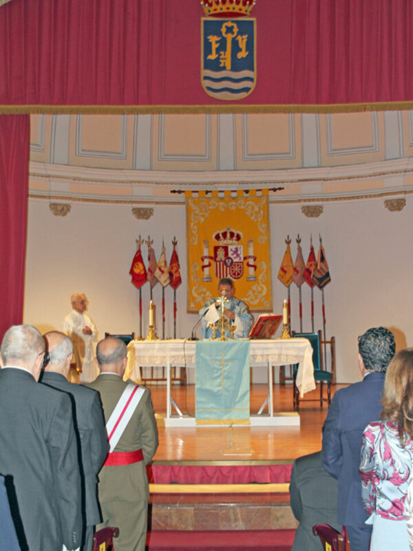 Ceremonia oficial con banderas y escudo real en el fondo, presidida por un sacerdote y asistida por autoridades en un salón formal.