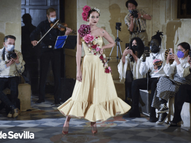 Model wearing a yellow dress with floral accessories, surrounded by photographers and musicians in a historic setting.