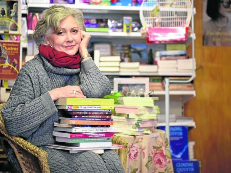 Una mujer sentada con una gran pila de libros en sus piernas, rodeada de estanterías de libros en un ambiente de librería.