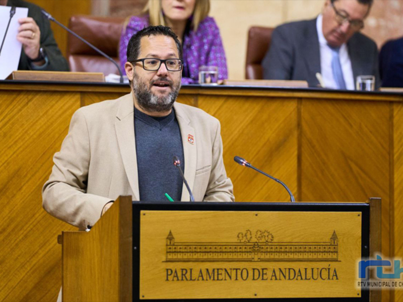 Un hombre hablando en un parlamento andaluz, con un fondo de madera y una placa con el nombre del parlamento en la parte inferior derecha.