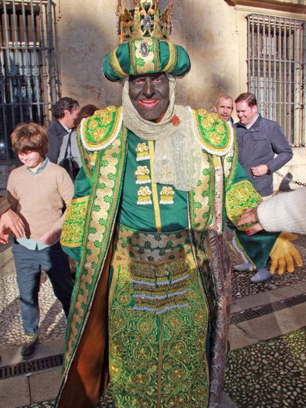 Personaje vestido como rey en traje verde y dorado, portando una corona y un bastón, en un entorno urbano.