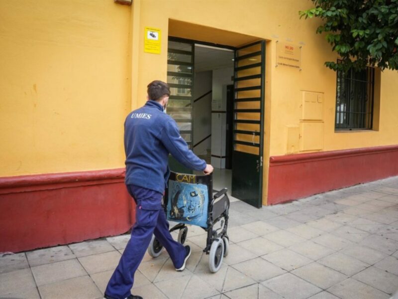 Persona con uniforme azul empujando una silla de ruedas frente a una entrada amarilla de un edificio.