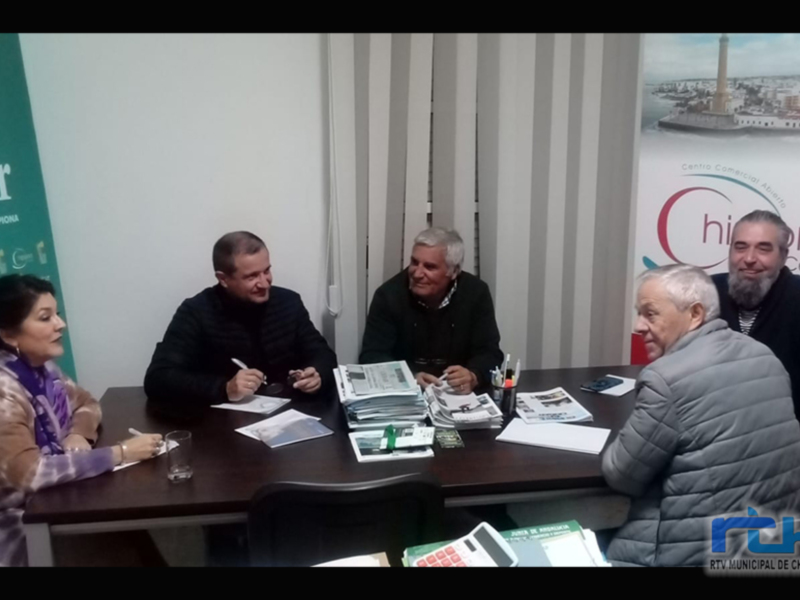 Un grupo de personas se reúne en una sala con un fondo que muestra una imagen de un puerto. La sala tiene una mesa con documentos y un banner con el nombre de un centro comercial.