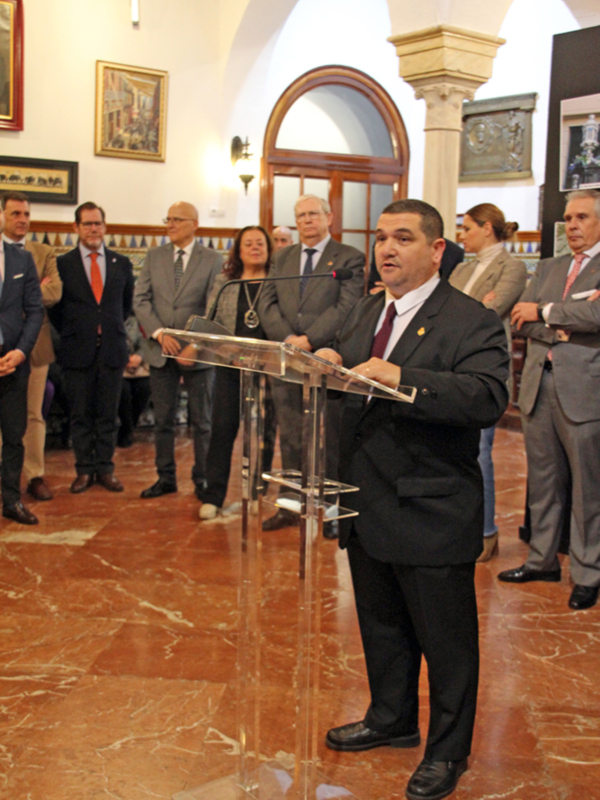 Persona hablando desde un podio en una sala con arcos y decoración clásica, rodeado por un grupo de personas en trajes.