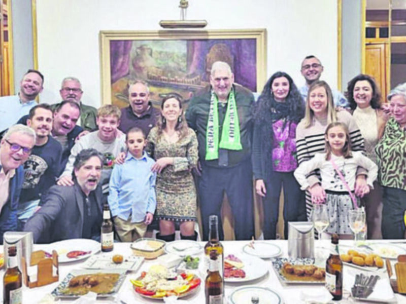 Celebración familiar en una mesa llena de comida y bebida, con un retrato colgante en el fondo.