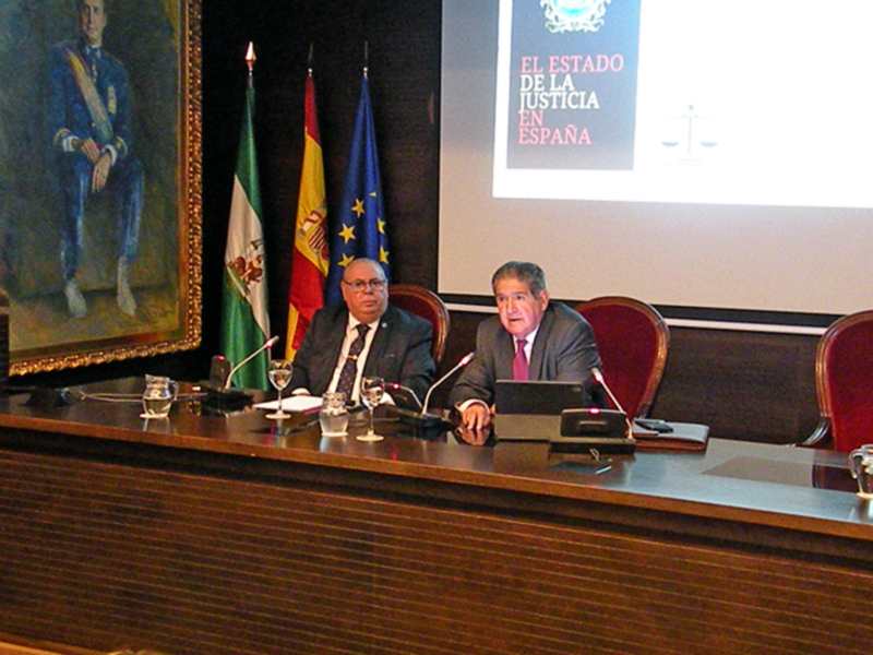Dos hombres en trajes se sientan en una mesa con bandejas de cristal, frente a una pantalla con el texto 'EL ESTADO DE LA JUSTICIA EN ESPAÑA'. Hay dos banderas, una española y una europea, y un retrato de un hombre en uniforme en el fondo.