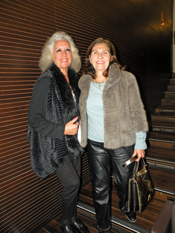 Dos mujeres sonríen y posan juntas en una escalera interior, vestidas con abrigos de piel y ropa formal, en un ambiente elegante.