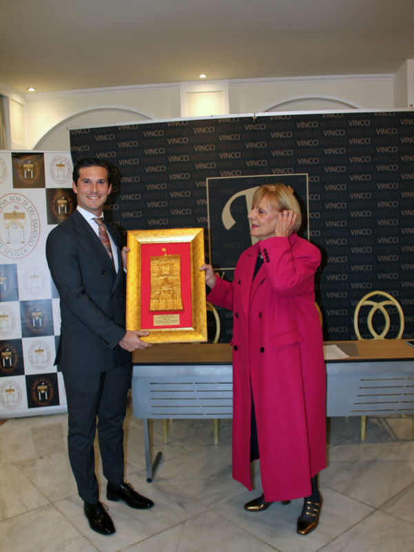 Persona con traje y mujer con abrigo rosa sostienen un trofeo dorado en un evento formal.