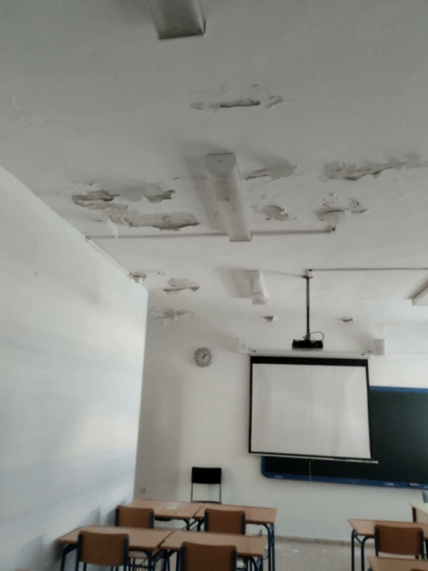 Salón de clase con pizarras y mesas vacías, mostrando señales de deterioro en las paredes y el techo.