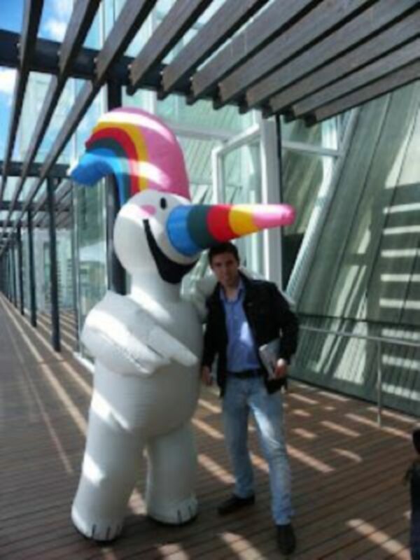Un hombre posa junto a un unicornio inflable con un arco iris en la cabeza, en un entorno moderno con estructuras de acero y vidrio. La imagen sugiere un evento o una celebración en un lugar público.