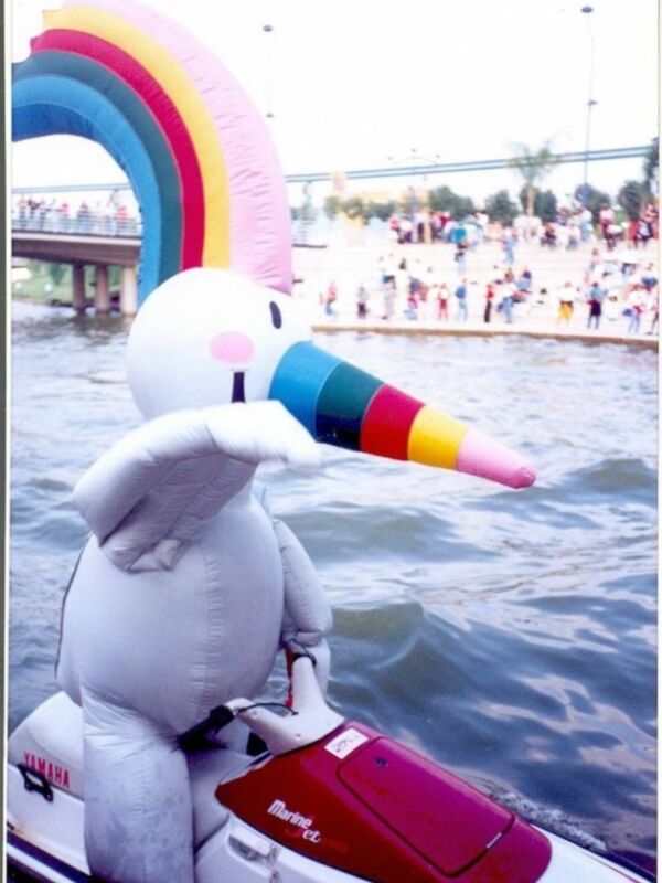Unicornio inflable en un barco en el agua, con un arcoíris en el fondo y una multitud de personas en la orilla.