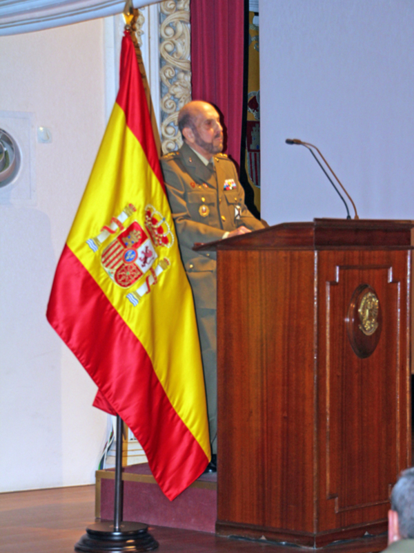 Oficial militar hablando en un evento formal con la bandera española en el fondo.