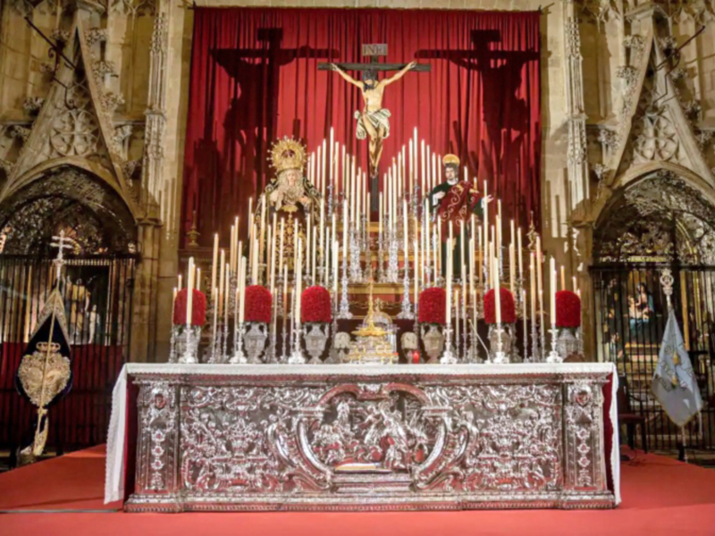 Altar con crucifijo y estatuas, rodeado de velas y decorado con mantel y alfombra roja en una iglesia gótica.