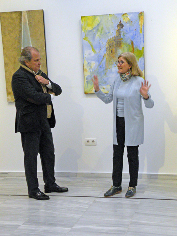 Dos personas interactúan en una exposición de arte, con una pintura de una torre en el fondo. La persona de la izquierda está ajustando su abrigo, mientras que la de la derecha se inclina hacia la pintura, posiblemente explicándole algo.