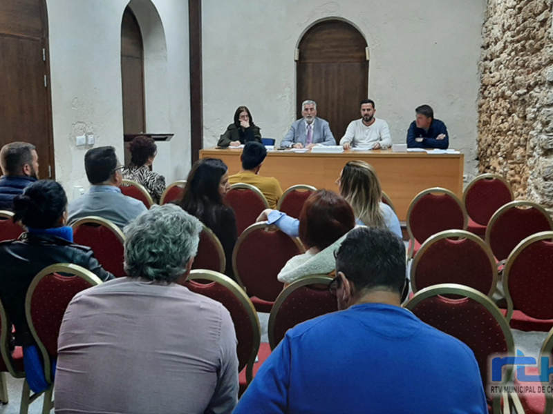 Sala de reuniones con personas sentadas en sillas rojas, un hombre en el centro hablando y otros asistentes escuchando.