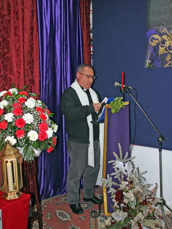 Un hombre en un vestido de oficio se dirige a un público, con un escenario decorado con flores y velas en el fondo.