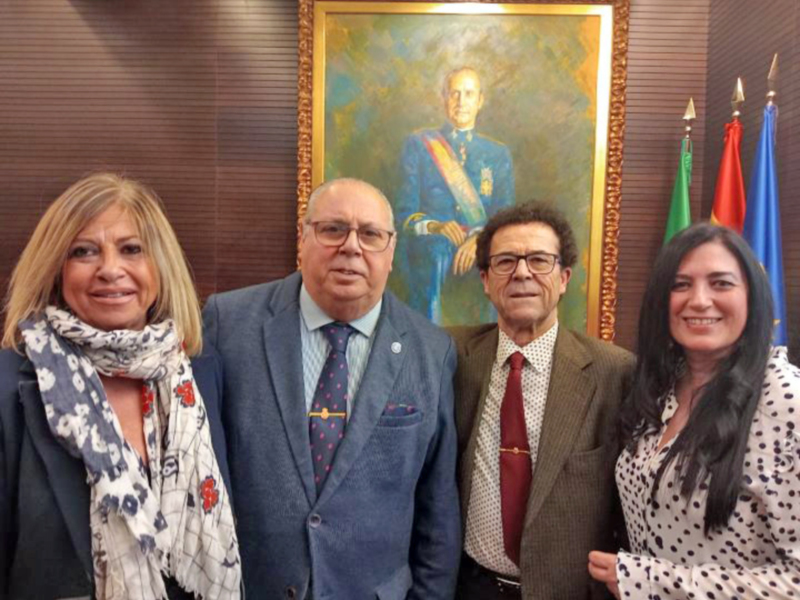 Tres personas posando frente a un retrato de un hombre vestido formalmente, con banderas en el fondo. La escena sugiere una reunión formal o una celebración oficial.