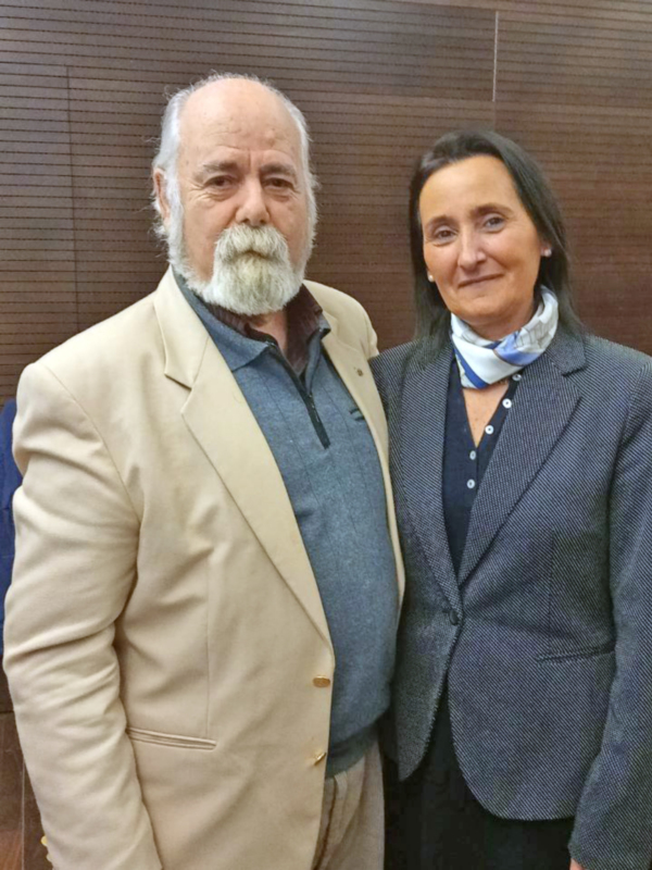 Dos personas vestidas formalmente posan juntas en un fondo de persianas.