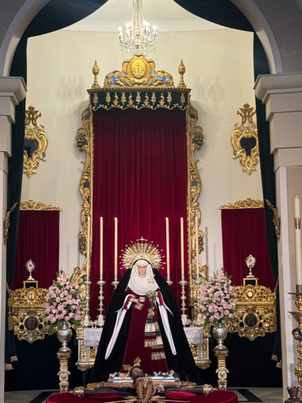 Altar con estatua de la Virgen María en trono dorado, rodeada de flores y candelabros, en un ambiente religioso formal.