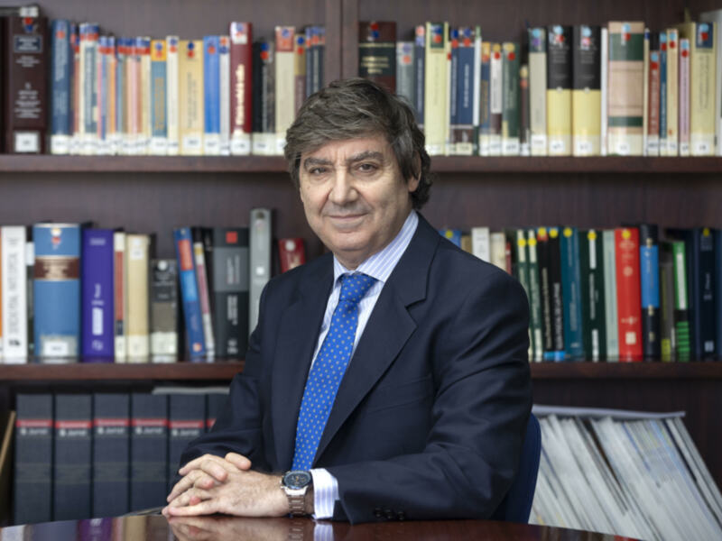 Profesional sentado en un escritorio con una estantería de libros de derecho detrás, vestido con traje y corbata azul.