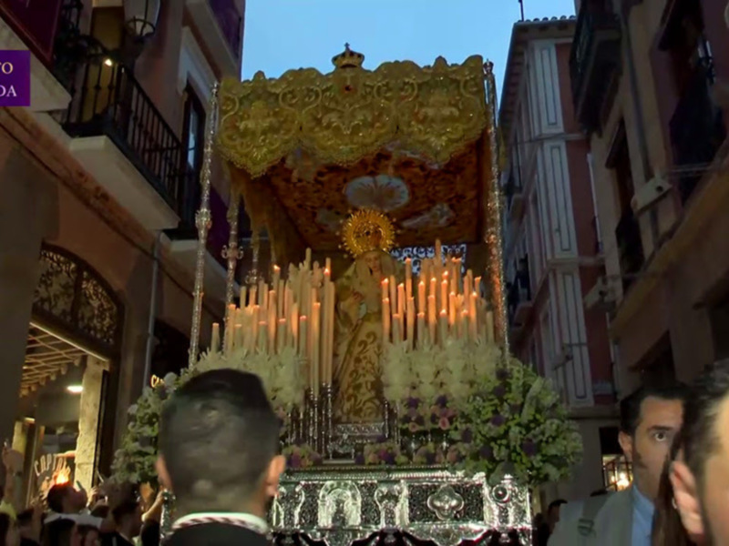Procesión religiosa en Granada con imagen de Virgen en un paso ornado con velas y flores.