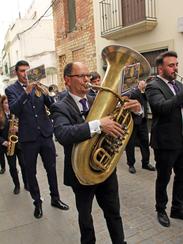 Banda de música tocando en una calle urbana, con músicos en trajes formales y instrumentos de viento y percusión.