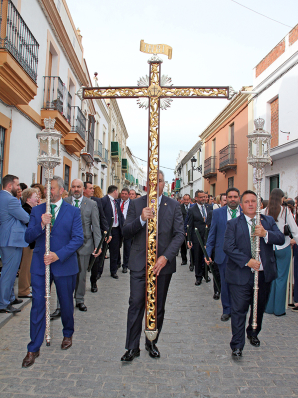 Procesión religiosa con hombres vestidos con trajes formales y una cruz dorada con inscripción INRI en la calle.