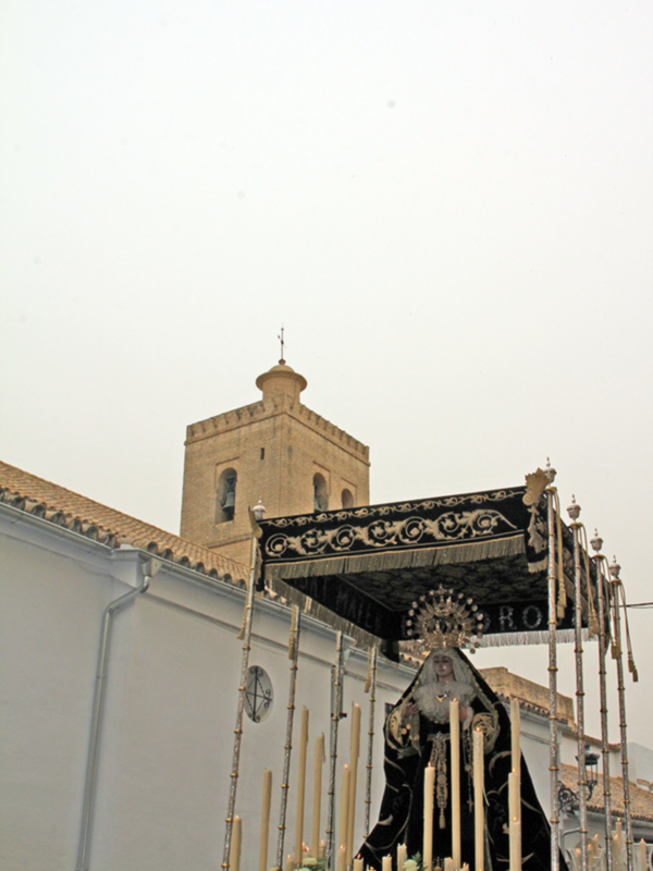Procesión con imagen de la Virgen de la Soledad en un día nublado, frente a una torre de iglesia.