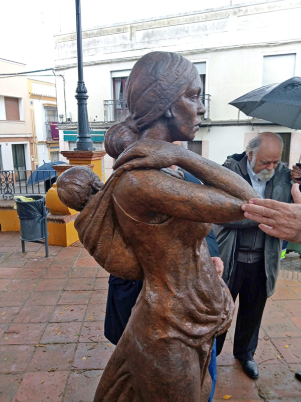 Escultura de bronce de una mujer cargando a un niño en su espalda, ubicada en una plaza urbana.