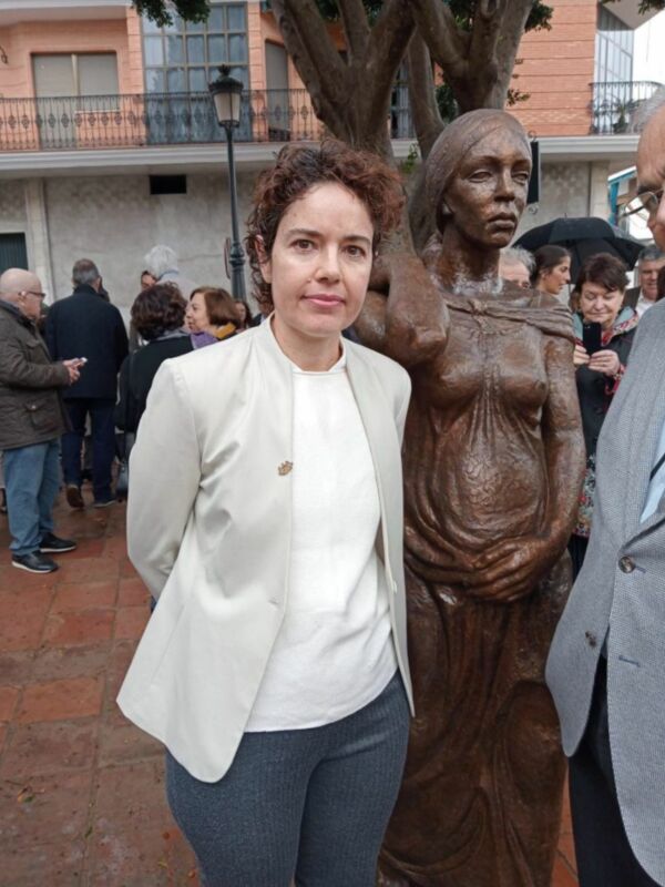 Persona posando junto a una escultura de mujer embarazada en un evento público al aire libre.