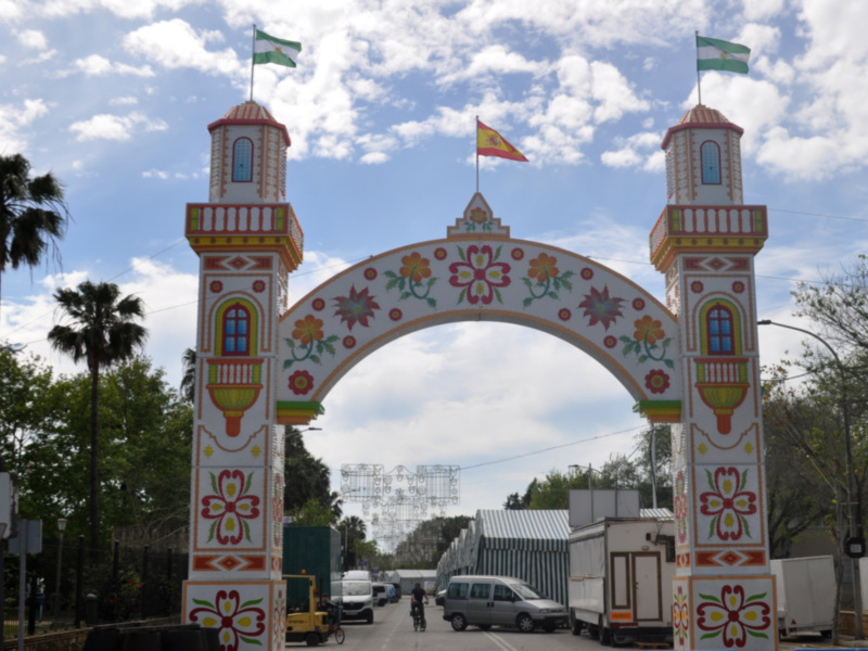 Arco decorativo con banderas y flores, típico de ferias andaluzas, bajo un cielo azul con nubes.