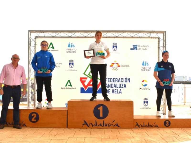 Premiación en el podio de una competición de vela, con tres participantes posando con sus trofeos y medallas, frente a un fondo con logos de organizaciones deportivas.