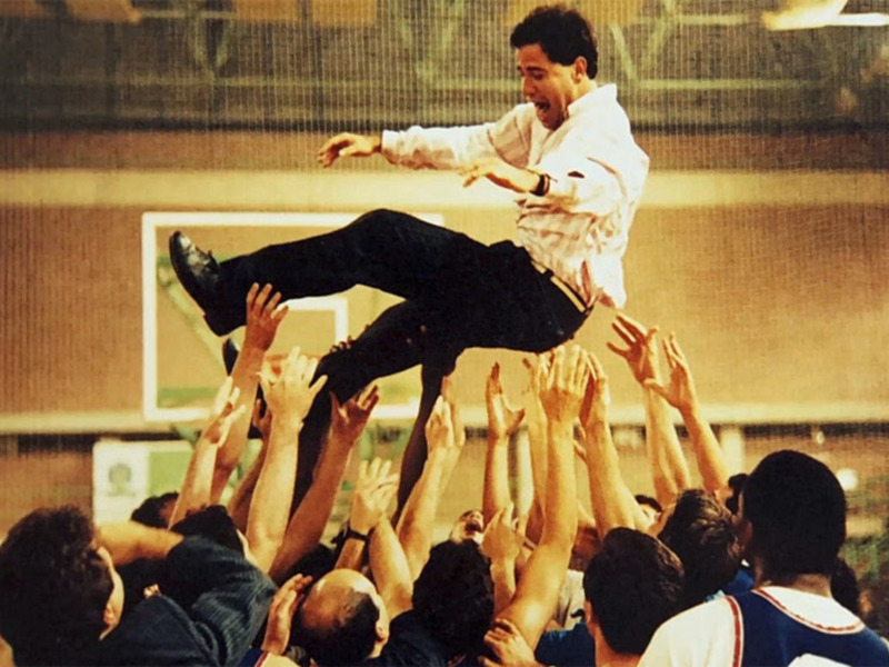 Un hombre en una camiseta blanca y pantalones negros está levantado por un grupo de personas en una cancha deportiva. La escena parece ser parte de una celebración o un evento deportivo.