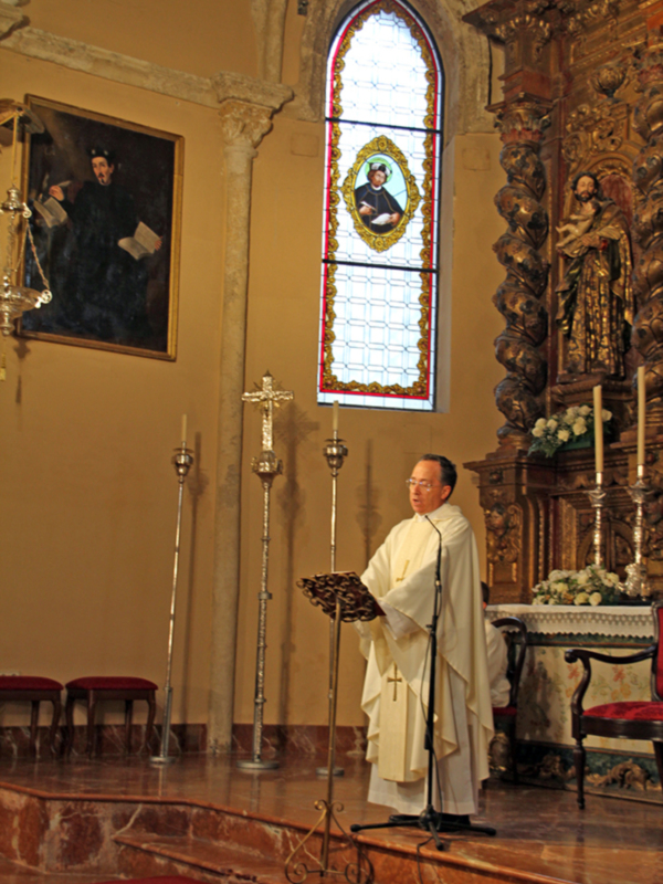 Misa en una iglesia con un sacerdote en vestido sacerdotal, un altar con flores, una vitrales con un retrato y un cuadro en la pared.