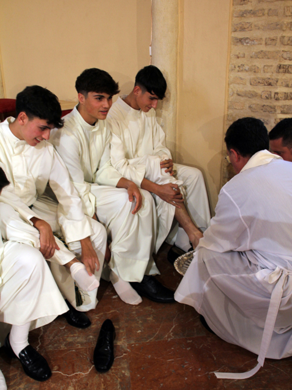 Jóvenes sacerdotes en traje blanco participan en una ceremonia religiosa, sentados en un ambiente con paredes de ladrillo.