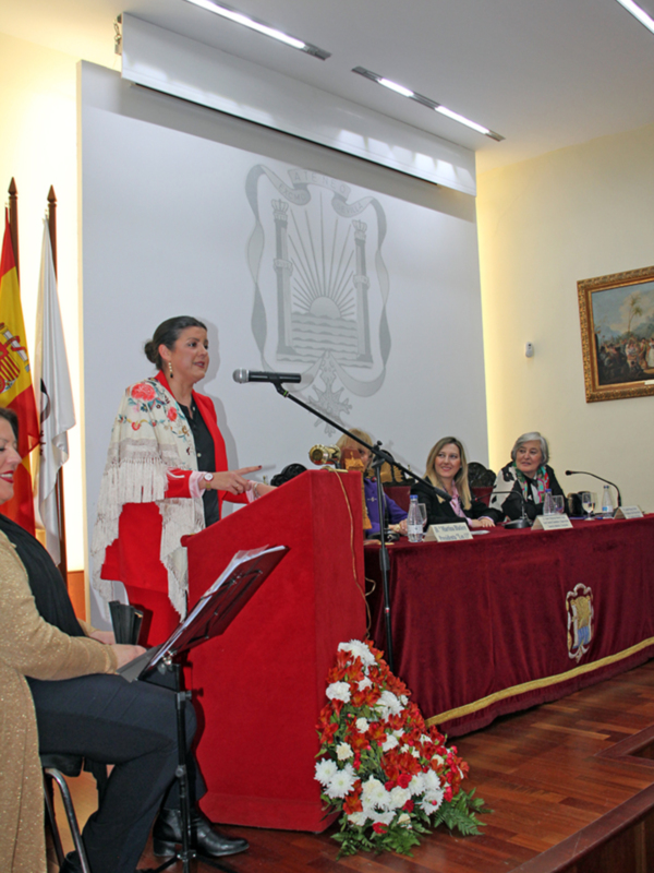 Una mujer hablando en un podium con un escudo nacional en el fondo, con una bandera española a su lado.