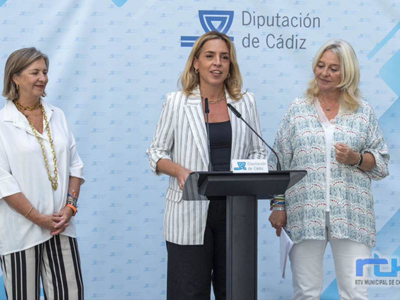 Tres mujeres en un evento público, con un fondo azul y blanco con el logo de la Diputación de Cádiz y el nombre de la localidad. La mujer en el centro está en un podio hablando, mientras que las otras dos la observan.