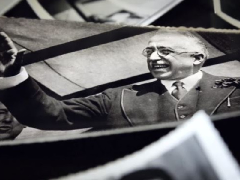 Retrato blanco y negro de un hombre con gafas levantando la mano, posiblemente en un acto público o político.