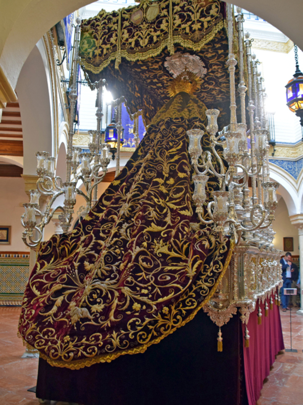 Procesión religiosa con manto bordado y candelabros dorados en un interior ornado.