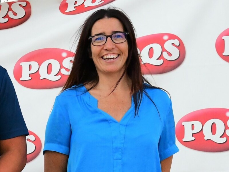 Una mujer sonriente con cabello largo y gafas, frente a un fondo con el logotipo de PQS.
