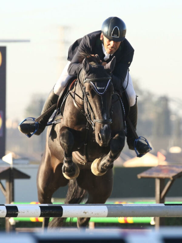 Un jinete monta un caballo en carrera, saltando sobre una barra de obstáculo. El jinete lleva un casco y ropa deportiva, mientras el caballo muestra su fuerza y agilidad. La escena se desarrolla en un entorno de competición, con una bandera visible al fondo.