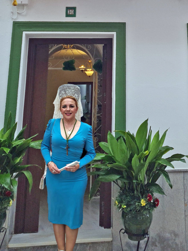 Una mujer con traje de fallera posa frente a una puerta decorada con detalles florales y plantas verdes.